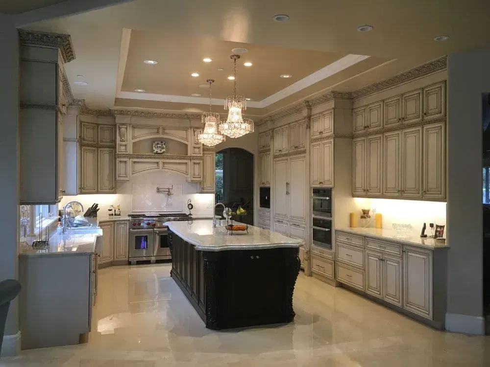 Open kitchen under a tray ceiling with paired crystal chandeliers above a dark island and glazed cream cabinetry