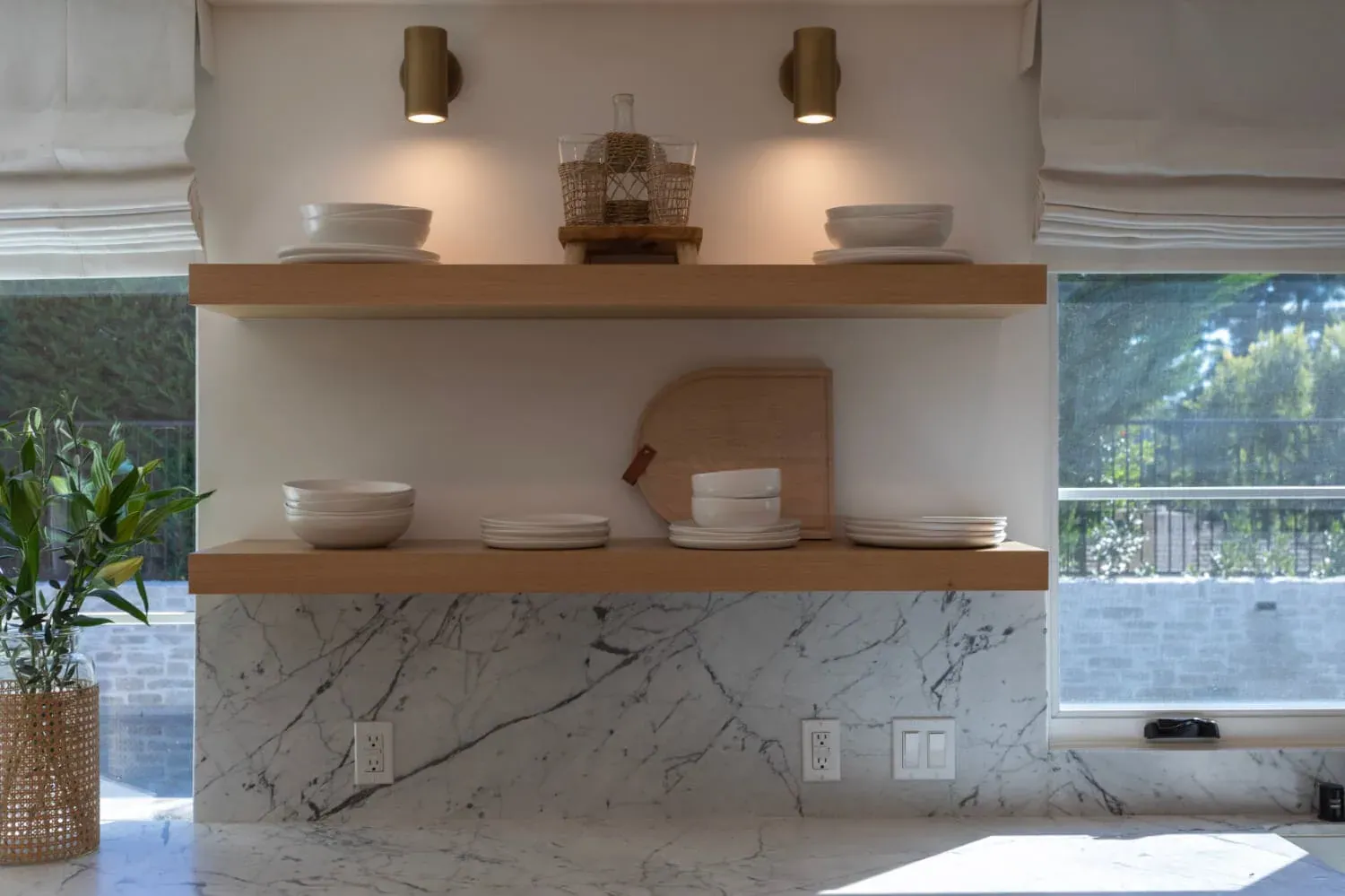 Marble slab backsplash behind floating oak shelves holding white dishes beside a window