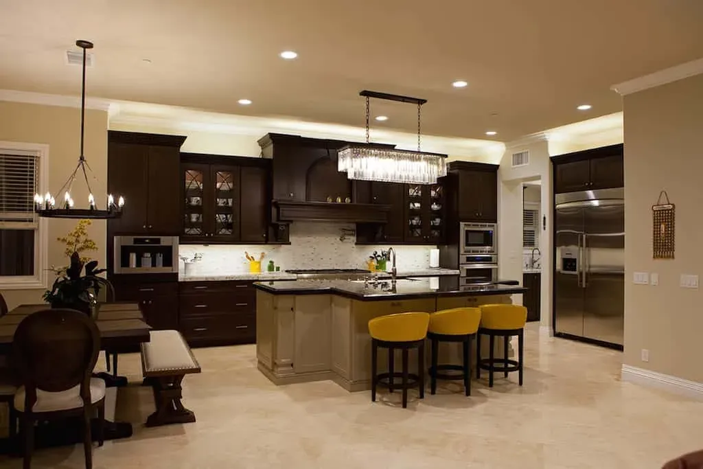Open-plan dark kitchen running into the dining area, with a linear crystal chandelier over the island and yellow bar stools