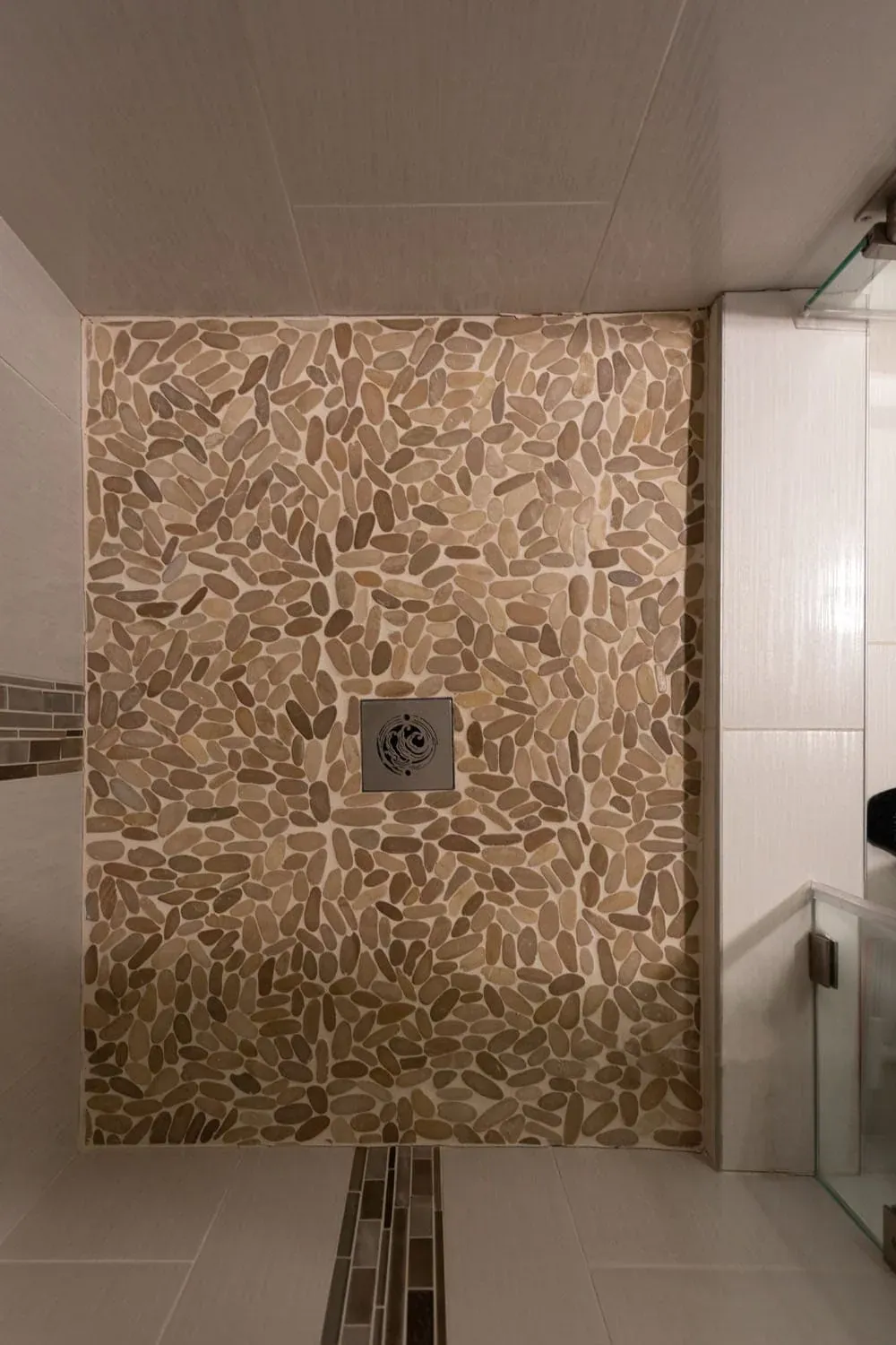 Shower floor laid in flat river pebbles around a square drain beside a white tiled wall
