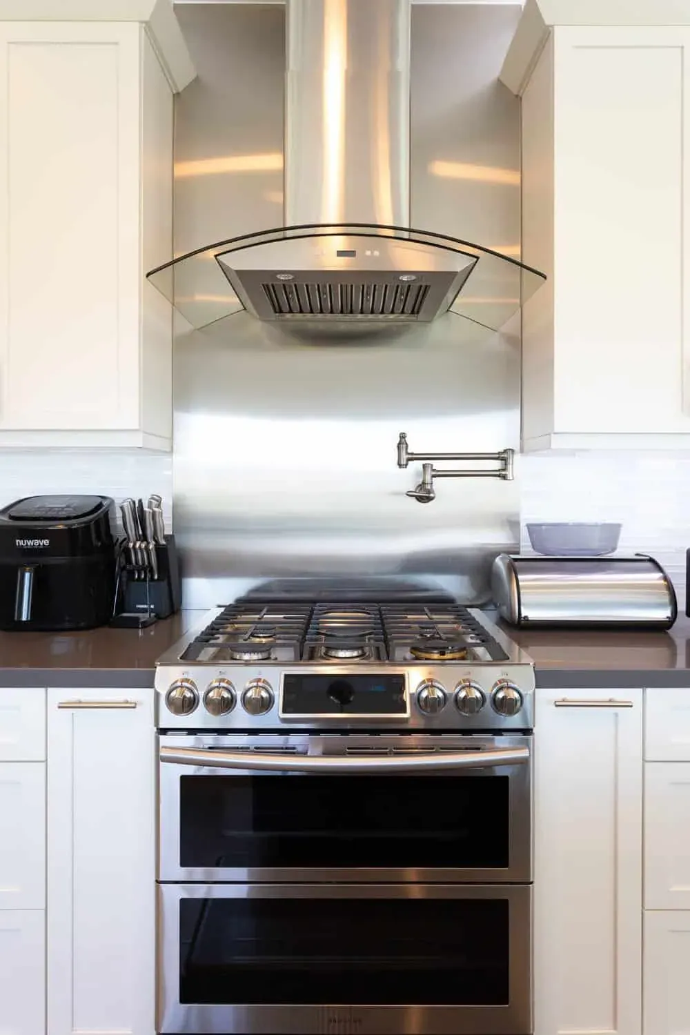 Stainless steel backsplash behind a gas range with a wall-mounted pot filler between white cabinets