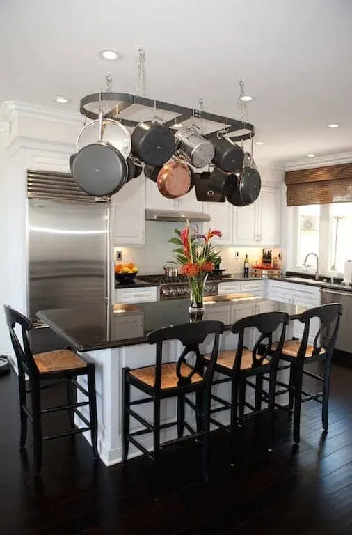 White kitchen with a hanging pot rack over a dark-topped island, rush-seat bar stools and a stainless refrigerator