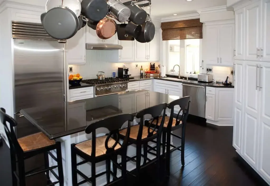 White kitchen cabinetry around a stainless refrigerator and range, with a pot rack over a dark island