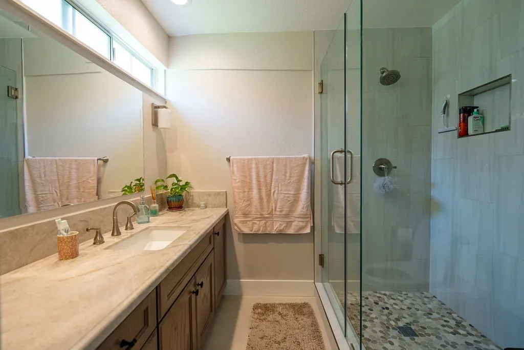 Bathroom with a stained oak vanity under a cream stone top beside a frameless glass shower with pale vertical tile