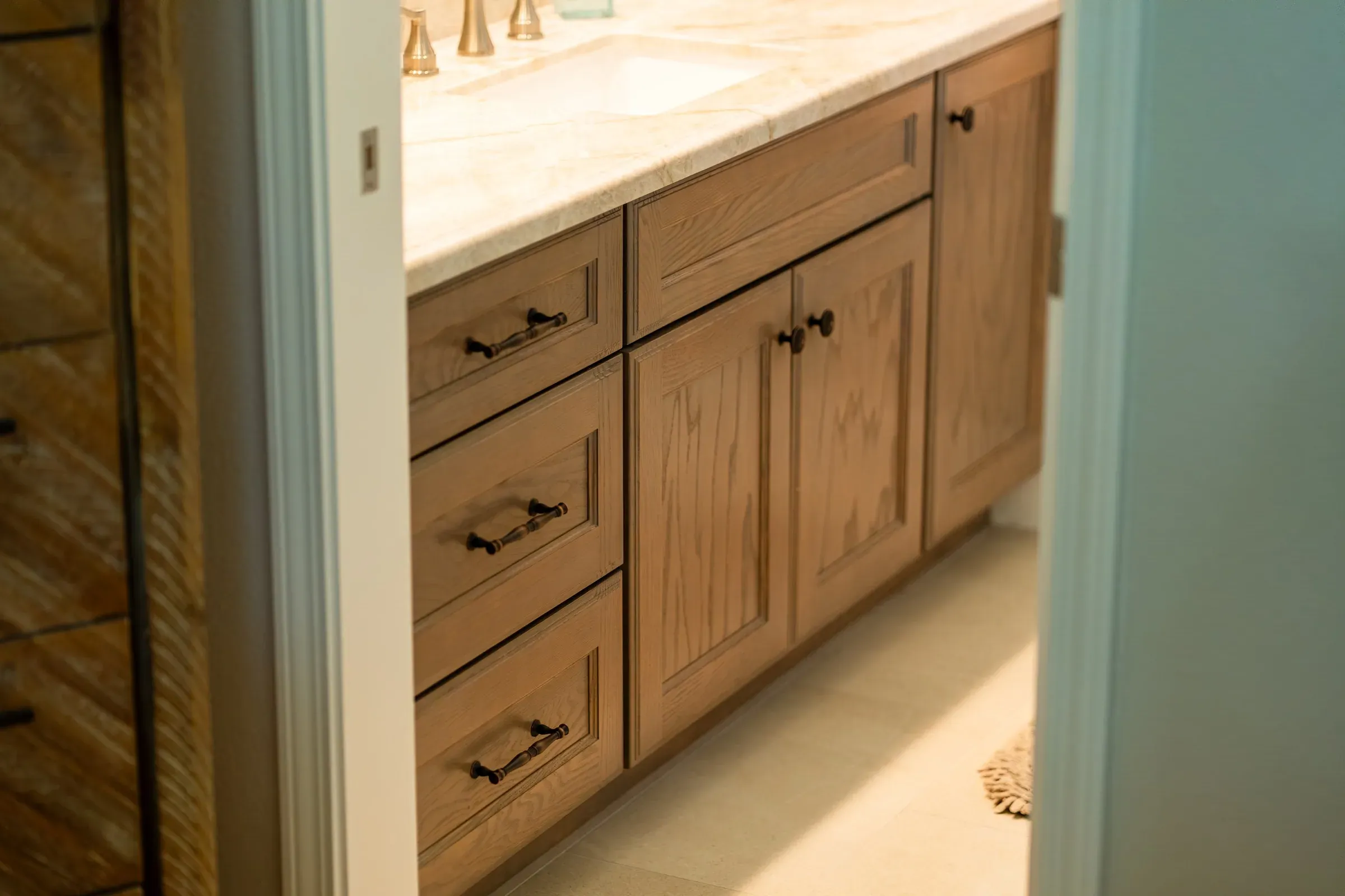 Stained oak bathroom vanity with a drawer bank and bronze pulls under a cream stone top, seen through a doorway