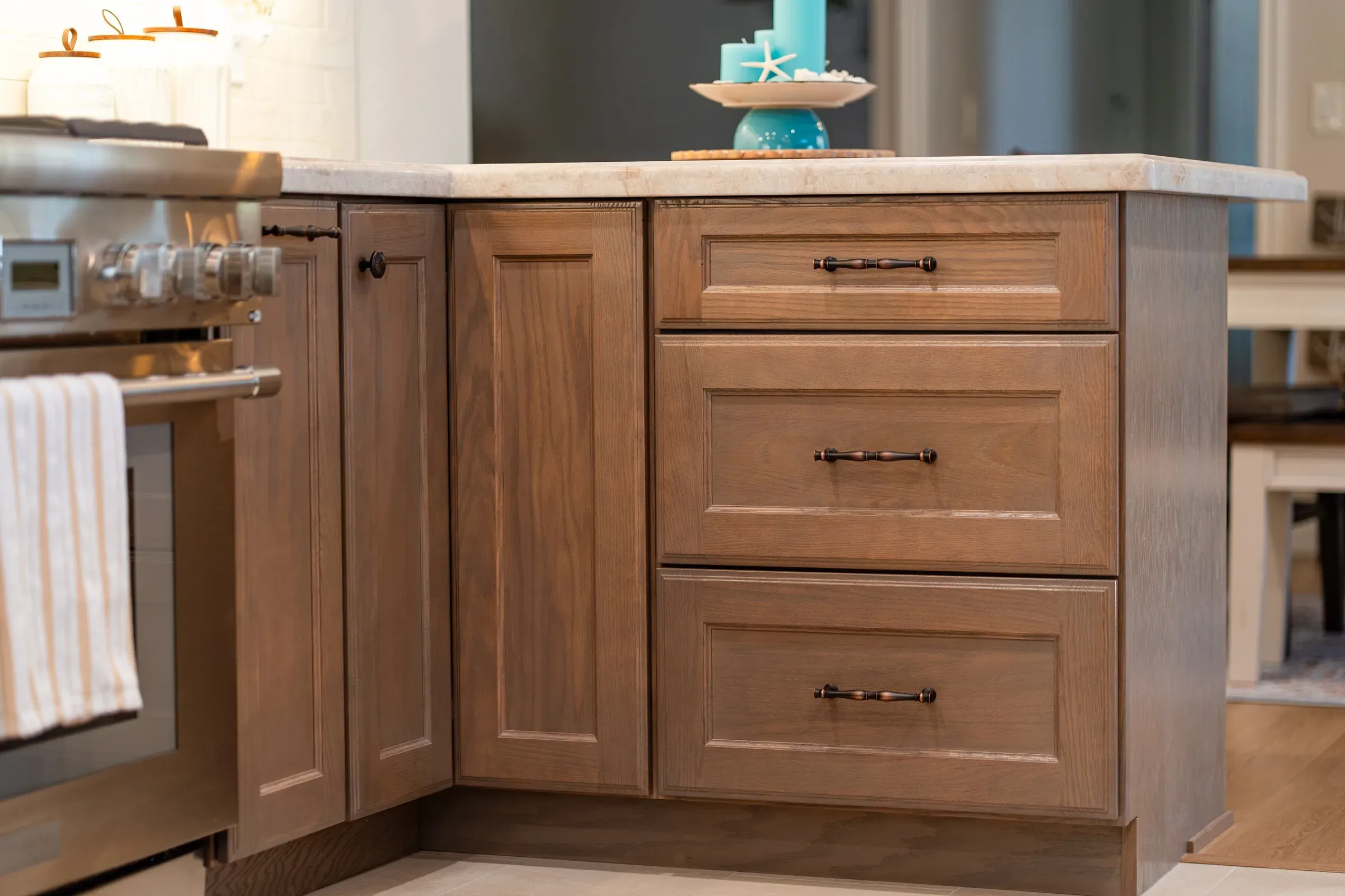 Stained oak drawer stack with bronze bar pulls at the end of a stone-topped run beside a stainless range