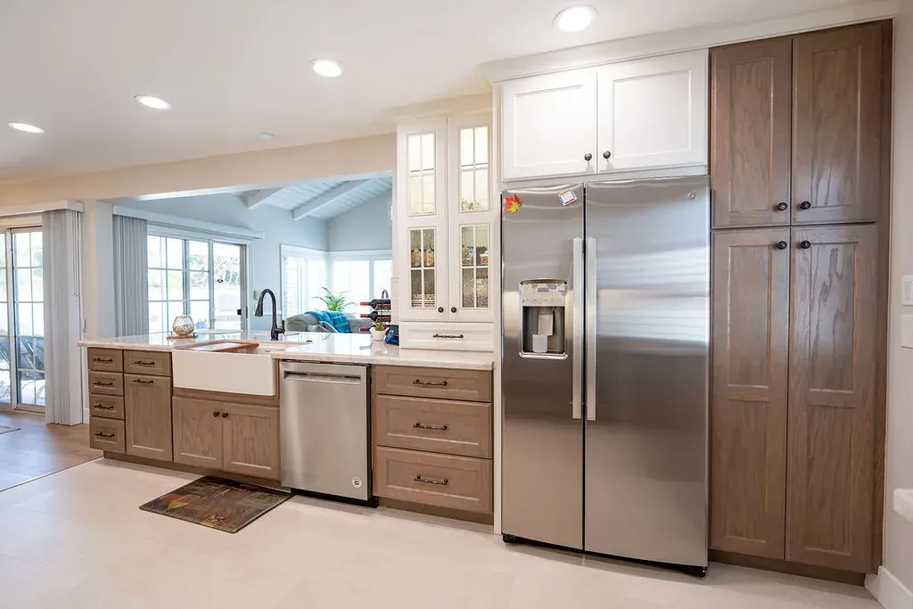 Stained oak pantry tower beside a stainless refrigerator, with white glass-front cabinets over the farmhouse sink run
