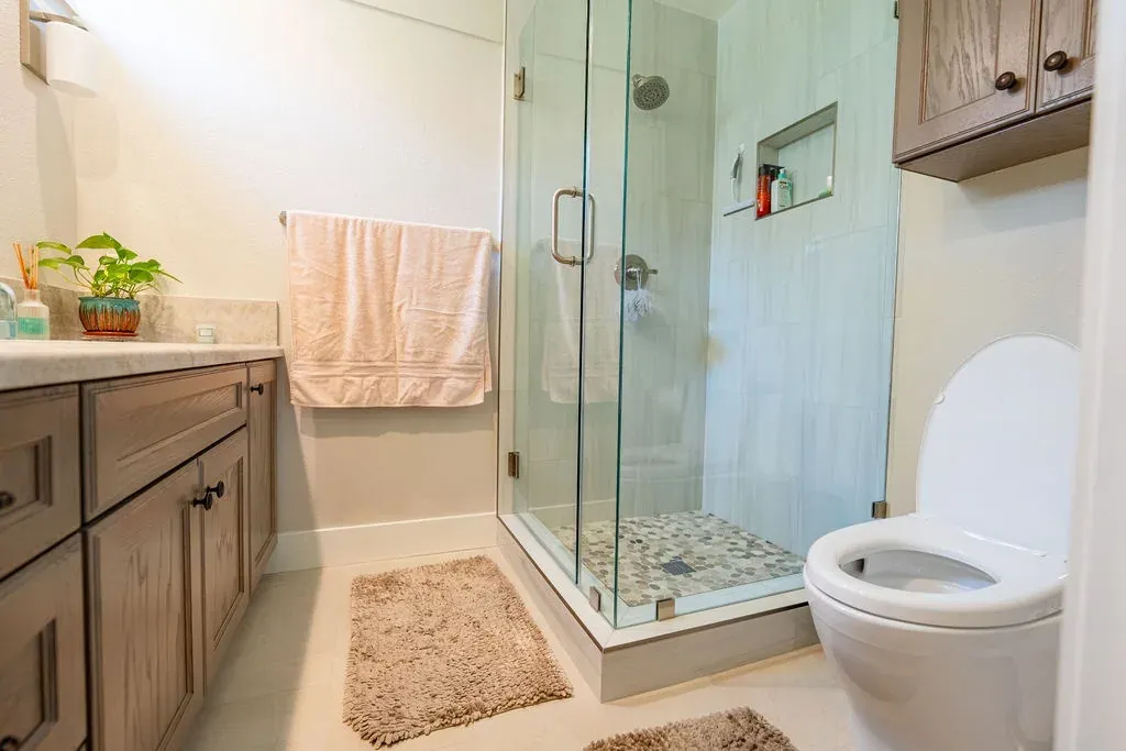Oak bathroom vanity with a stone top next to a frameless glass shower with a pebble floor, under an oak wall cabinet