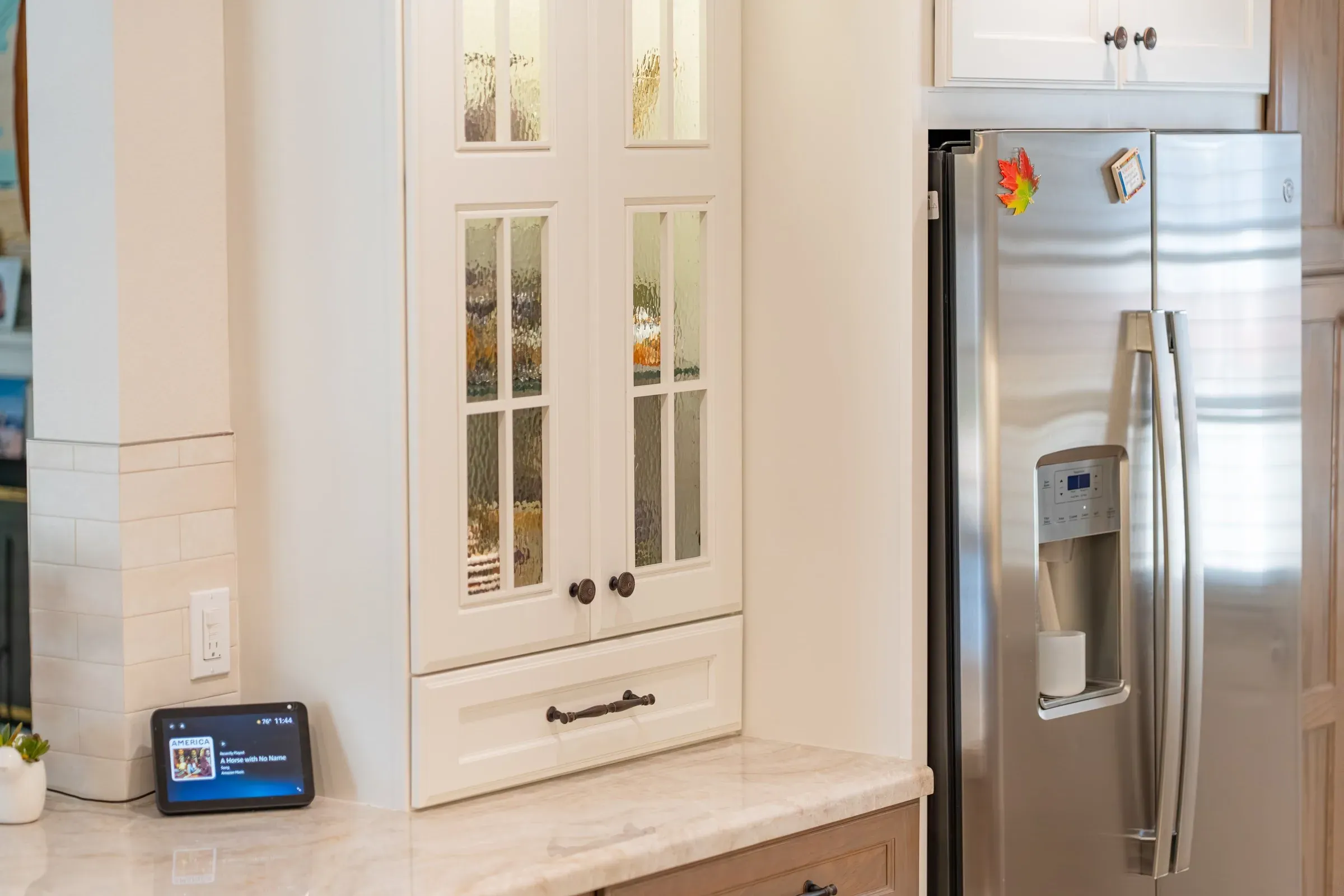 Tall white glass-front cabinet beside a stainless refrigerator above a tiled counter