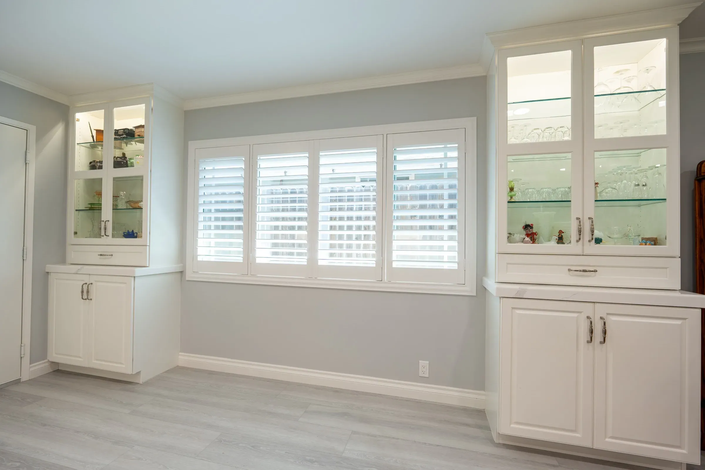 Two white glass-front hutch cabinets with lit glass shelves flanking a window fitted with white plantation shutters