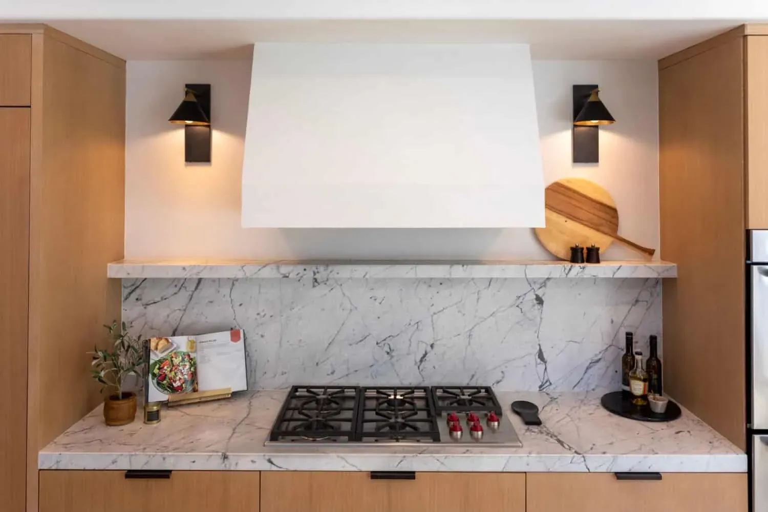 White plaster range hood between two wall sconces above a gas cooktop and a marble slab backsplash