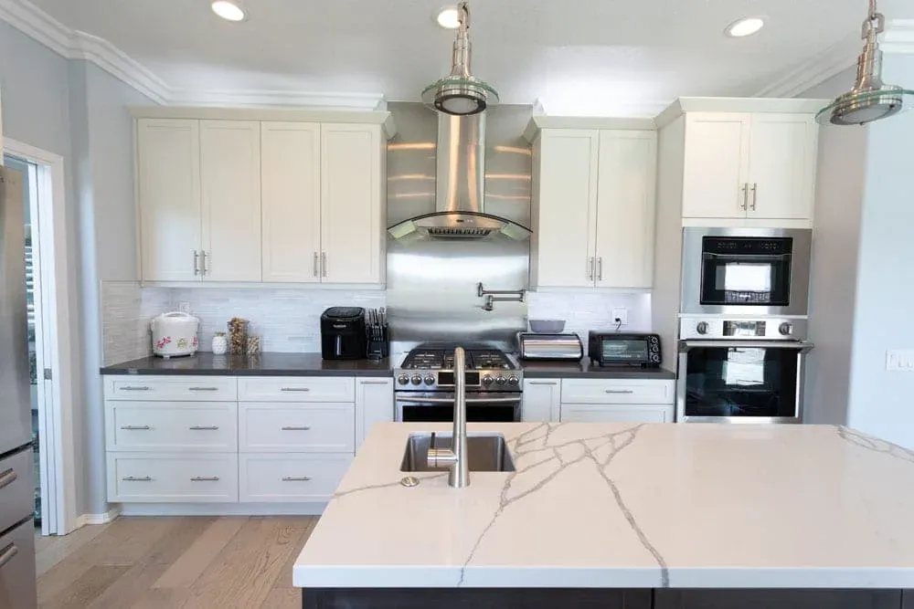 White shaker kitchen with a stainless chimney hood, wall ovens and a veined island