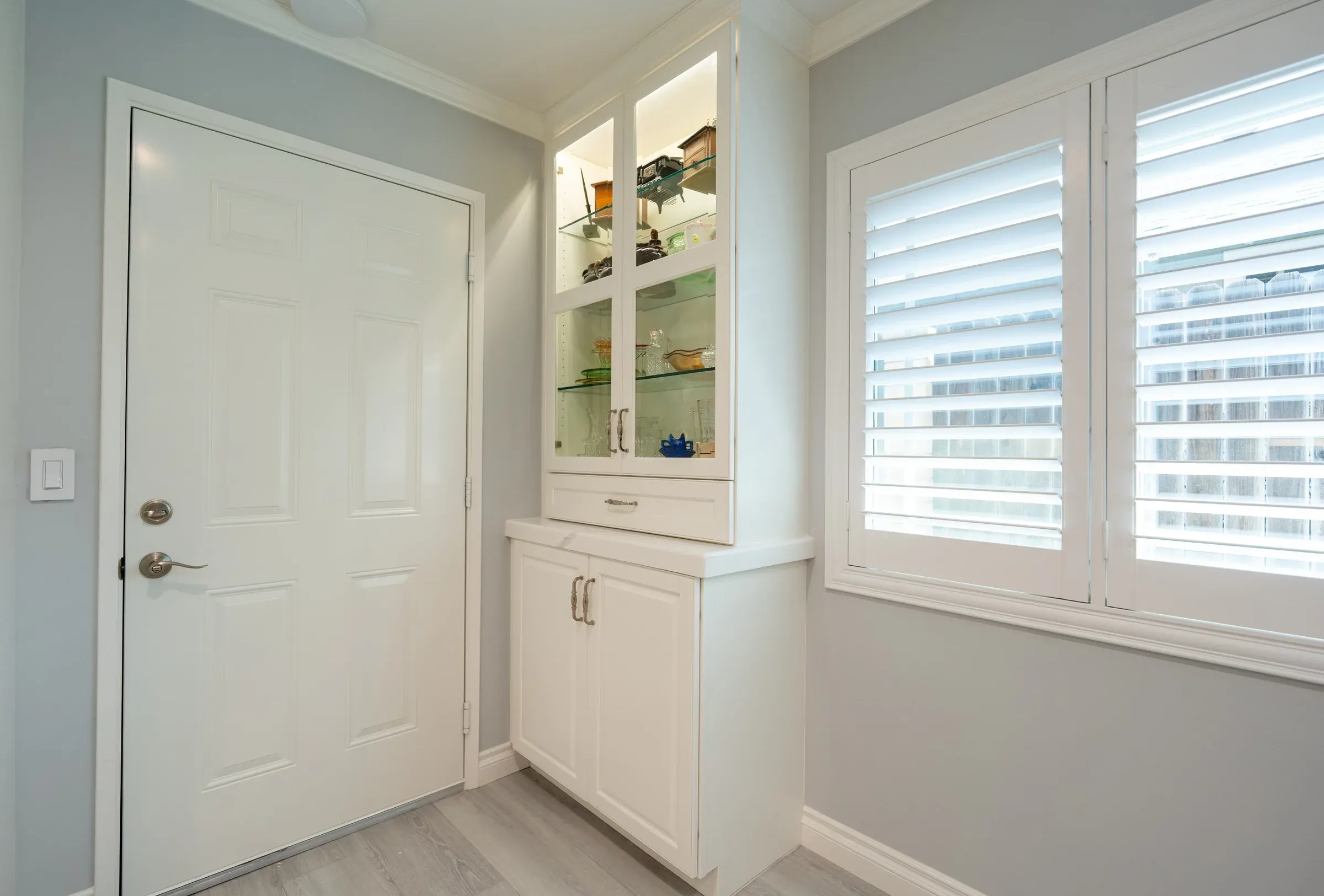 Tall white cabinet with lit glass-front doors beside a window with white plantation shutters