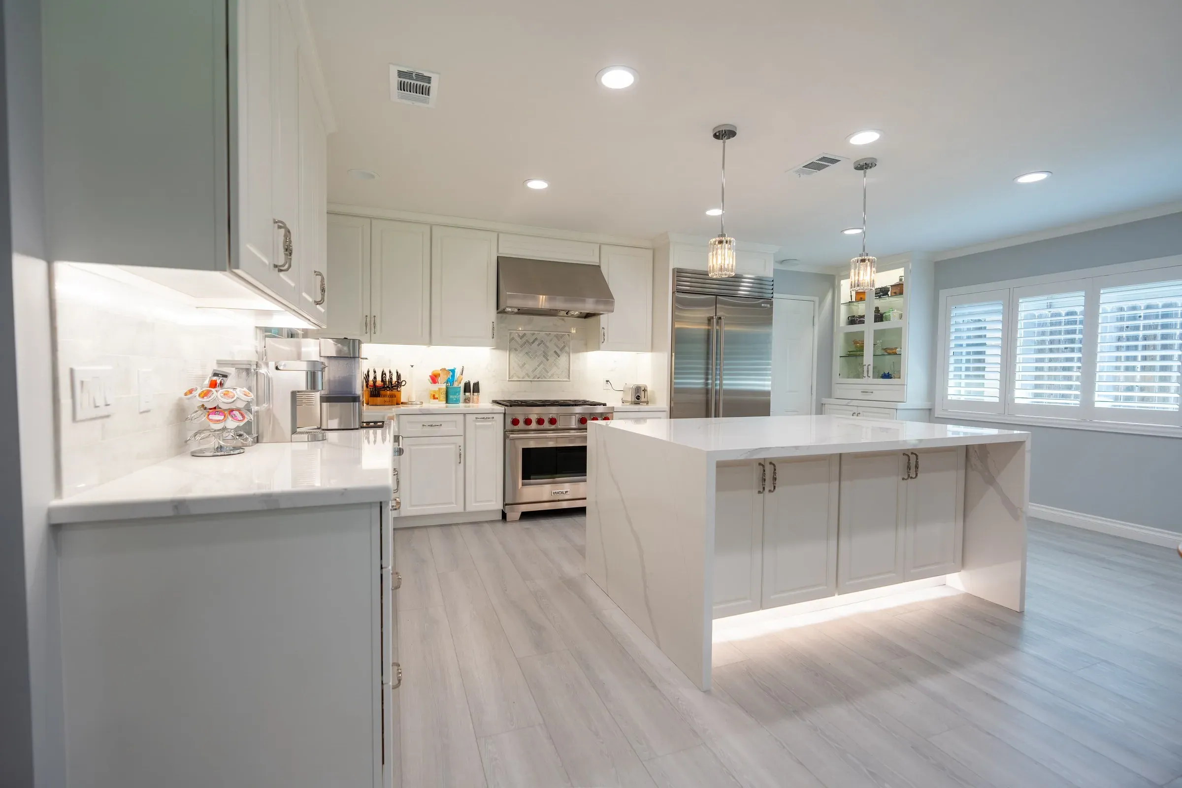 White raised-panel kitchen with a quartz waterfall island lit along its base and a stainless range with red knobs beyond